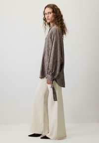 Femeie cu păr creț, purtând o bluză maro cu un model lejer, pantaloni crem cu picioare largi și tivuri destul de tocite, stând de profil.