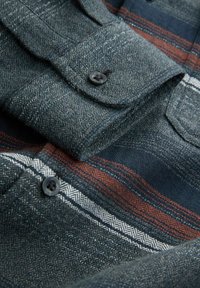 Rakstainā flanelēta krekla tumši zilā pamata krāsa ar pelēku tekstūru un akcentējošām svītrām sarkanā un baltā krāsā. Ietver pogājamu apkakli un kabatas.