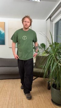 Zelena kratka majica s kratkimi rokavi in okroglim logotipom, črne hlače in sive čevlje. Notranji prostor z rastlinami in vidnim sivim kavčem.