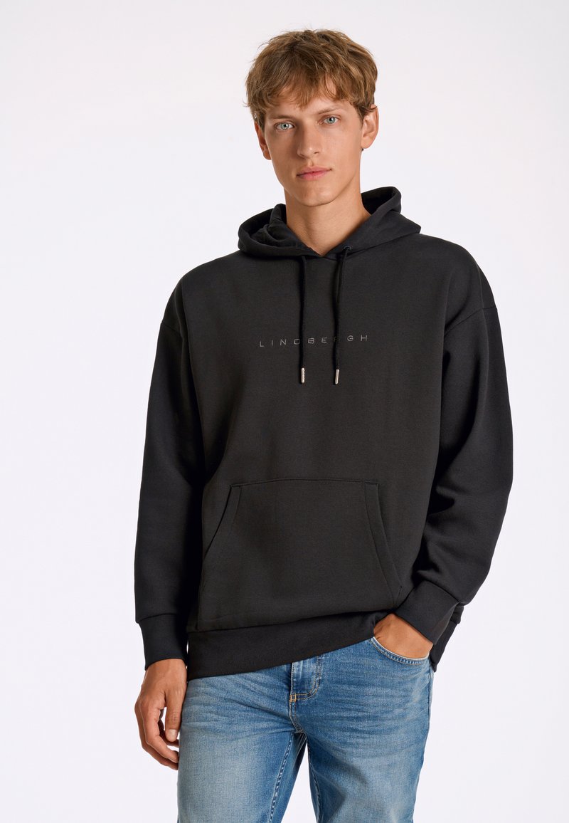 Sudadera negra con capucha, bolsillo delantero, cordones y estampado de logo. Combinada con vaqueros de un azul claro; tejido suave y texturizado.