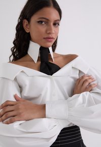 Camicia bianca con colletto drammatico a spalle scoperte e maniche ampie, abbinata a un accento di cravatta nera. Presenta dettagli ricamati sui polsini.