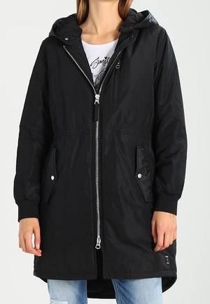 Kobieta ubrana w czarną, długą kurtkę z kapturem, zapinaną na zamek i zatrzaski, noszoną na białej koszulce z grafiką oraz niebieskich dżinsach.