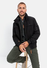 Black insulated jacket with snap buttons and zippered pockets, worn over a gray sweater. Green pants and light sneakers complete the look.