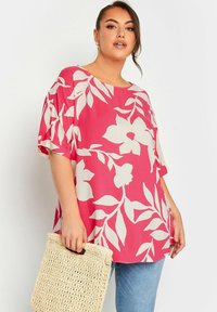 Blusa rosa larga con grande motivo floreale bianco, maniche corte, scollatura tonda, abbinata a una borsa di paglia beige.