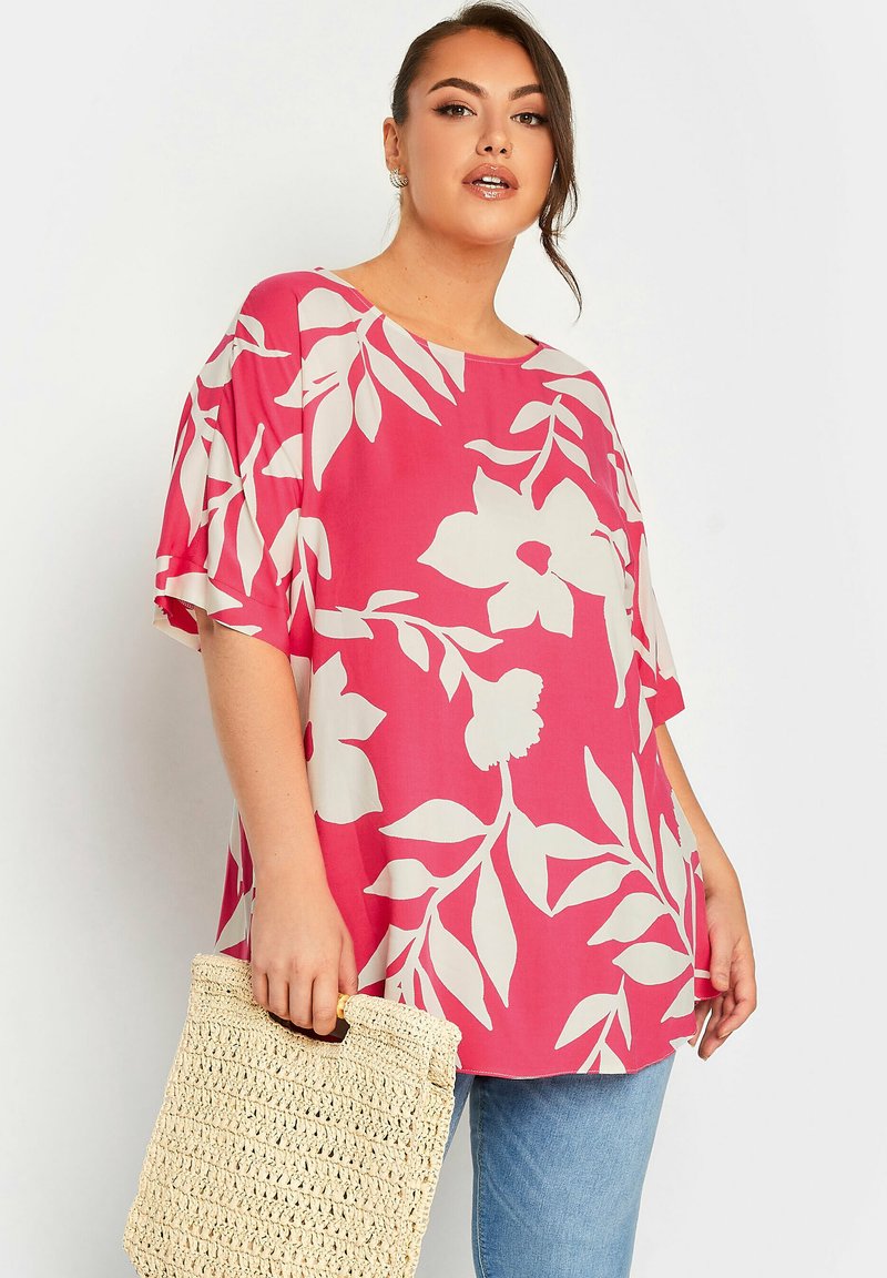 Blusa rosa larga con grande motivo floreale bianco, maniche corte, scollatura tonda, abbinata a una borsa di paglia beige.