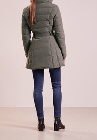 Olivgrön puffjacka med huva, åtdragen midja och vertikal quiltad design, tillsammans med mörk denimjeans och svarta ankelstövlar.