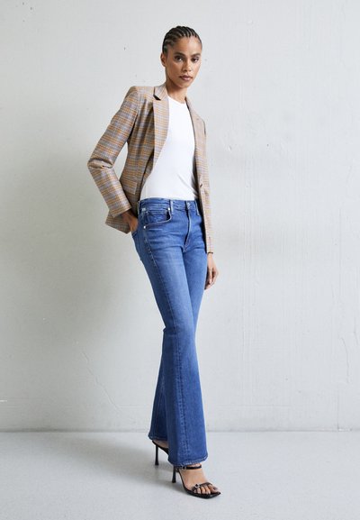 Blazer a quadri in marrone e beige sopra un top bianco aderente, abbinato a jeans a campana blu e tacchi neri con cinturini. Superfici texture e vestibilità sartoriale.