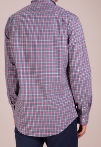 Kofta med långa ärmar i bomull, i ett flerfärgat rutmönster med nyanser av rosa, blått och vitt; har en rundad nederkant och knäppningar vid ärmarna.