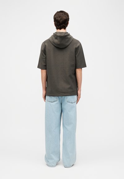 Άνδρας που φοράει σκούρο κοντομάνικο φούτερ με κουκούλα και ανοιχτομπλε φαρδιά τζιν παντελόνια, στέκεται με την πλάτη προς τα εμάς μπροστά σε απλό ανοιχτό φόντο.