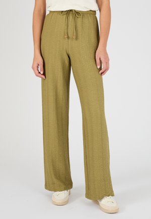 Mujer con pantalones anchos de pierna ancha color verde oliva con textura y cintura con cordón, y zapatillas blancas sobre un fondo claro y liso.