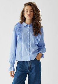 Blusa a righe azzurro chiaro con dettagli arricciati sulle spalle, chiusura con bottoni e polsini arricciati, abbinata a jeans in denim.