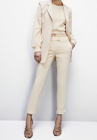 Beige skräddarsydd kavaj över en matchande kortad tröja och slim-fit byxor. Outfiten har en mjuk textur och en minimalistisk design. Beiga klackar med remmar fulländar looken.