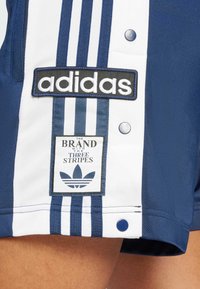 Tamnoplave atletske kratke hlače s bijelim prugama, s izvezenom "adidas" oznakom i etiketu s natpisom "Brend s tri pruge."