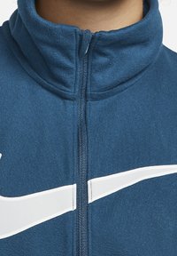 Mørkeblå lynlåstrøje med høj krave, hvid logoaccent og glat stoftekstur med en struktureret pasform.