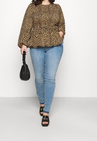 Frau in einer Bluse mit Leopardenmuster, hellblauen Skinny-Jeans, schwarzen Sandalen, die eine kleine schwarze geraffte Handtasche hält und nach vorne geht.