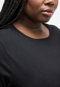 Κοντινό πλάνο ενός ατόμου με πλεξούδες, που φοράει απλό μαύρο μπλουζάκι με στρογγυλή λαιμόκοψη, εμφανίζοντας μόνο τα χείλη και το κάτω μέρος του προσώπου.