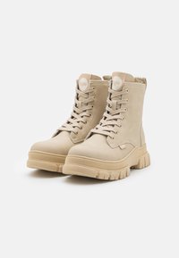 Botas de combate bege, feitas de couro e têxtil, com colarinho almofadado, sola de borracha espessa e alça lateral para fácil calce.
