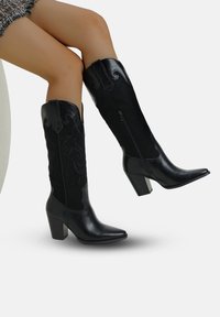 Botas negras hasta la rodilla con punta afilada, que combinan cuero y ante, con intrincados patrones grabados y un tacón ancho.