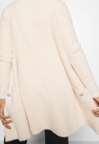 Krémově zbarvený oversize kardigan z jemného texturovaného materiálu s bočními kapsami zdobenými knoflíky a vroubkovaným lemem.