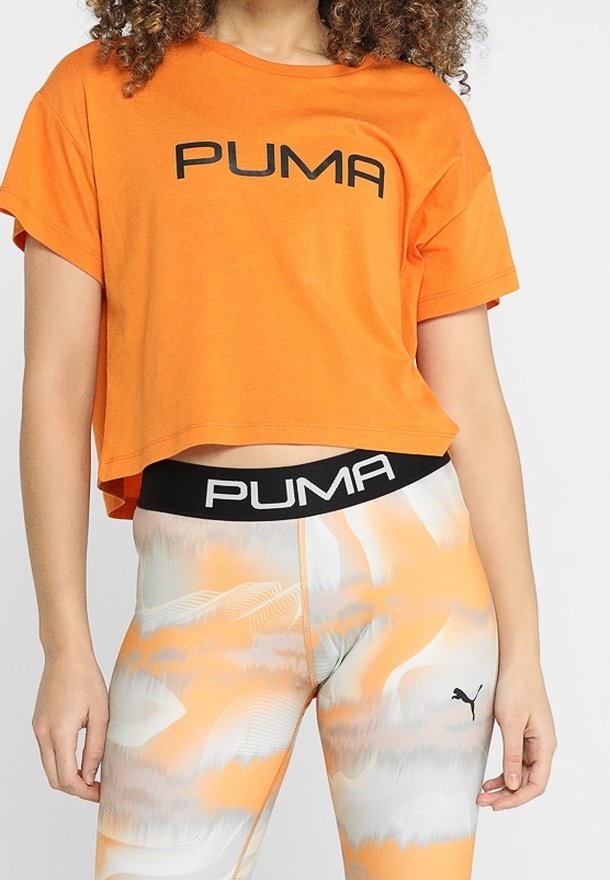 Femme portant un crop top orange avec le texte "PUMA" et des leggings assortis orange et blanc à motif avec le logo "PUMA" et une ceinture noire.