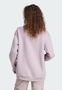 Ljuslila sweatshirt med texturerat mönster, sänkta axlar, ribbade muddar och nederkant. Utsikt från baksidan som visar lockigt hår.