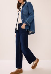 Mujer con zapatillas marrones, jeans de un azul oscuro, una camiseta blanca y una chaqueta de mezclilla azul holgada, de pie con una mano en el bolsillo.