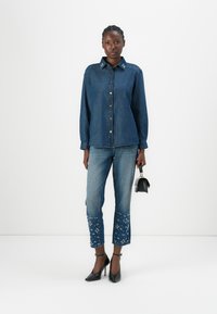Cămașă din denim cu un nasture în față și guler brodat, asociată cu blugi albaștri deschis cu broderie florală la tiv și tocuri negre.