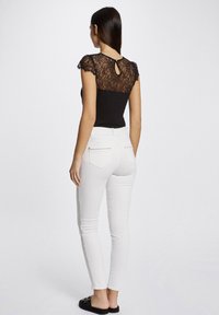 Top en dentelle noire avec un dos en keyhole, associé à un jean skinny blanc. La tenue présente des détails subtils et une silhouette ajustée.