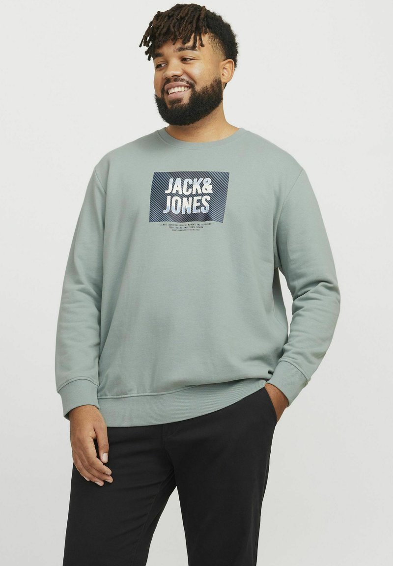 Sudadera verde claro con cuello redondo y puños de canalé. Presenta un logotipo impreso en azul marino y blanco. Combinada con pantalones negros.