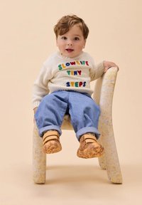 Un niño pequeño con cabello rizado está sentado en una silla pequeña, vistiendo un suéter blanco con texto colorido, jeans azules y zapatos marrones.