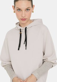 Felpa leggera beige con lacci neri, dotata di accenti ribassati metallici argento sulle maniche. Tessuto morbido, vestibilità rilassata.