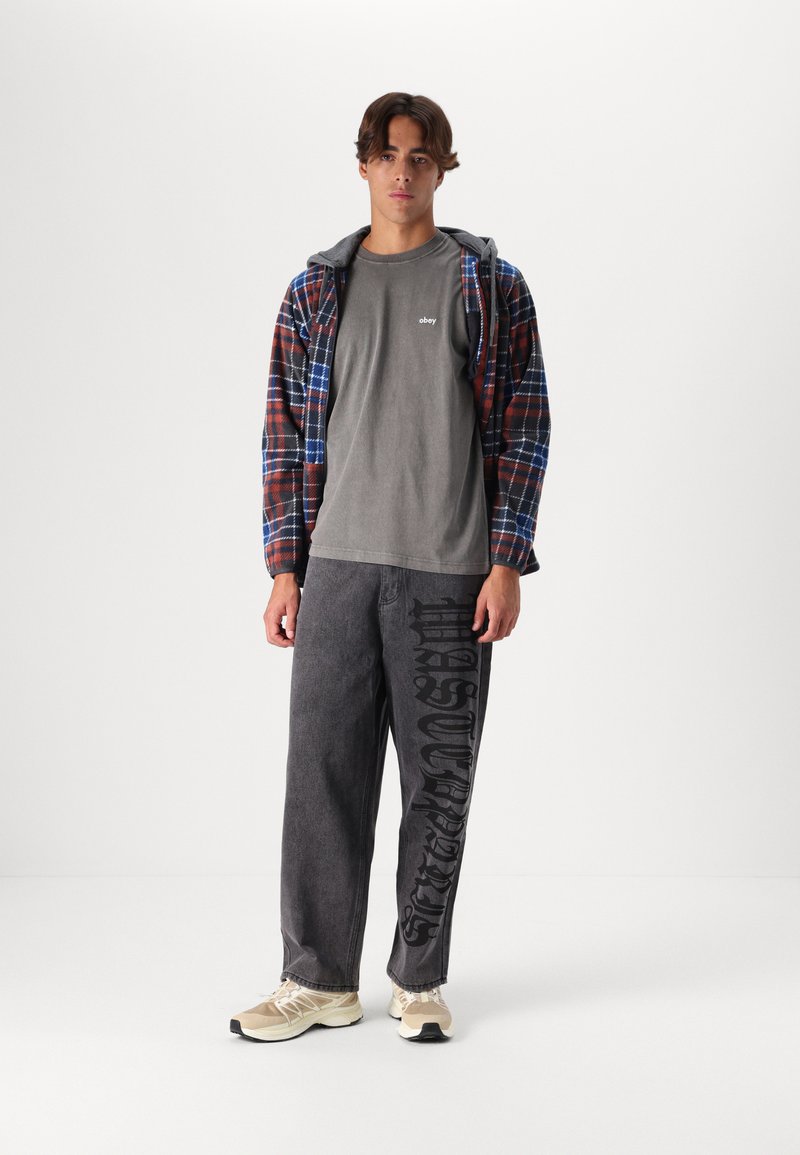 Plaid flannel jacket in red and blue, gray t-shirt with logo, gray baggy pants with black text, and beige athletic shoes.