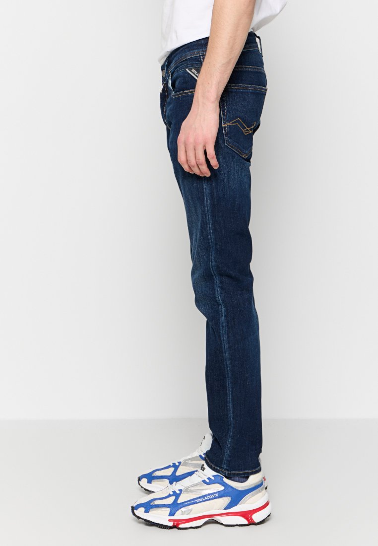 Jeans slim-fit blu scuro con cuciture a contrasto, cinque tasche e design a gamba dritta, abbinati a sneakers bianche con accenti blu e rossi.