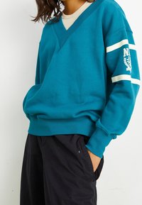 Teal v-ringad sweatshirt med långa ärmar, med krämfärgade ränder och logotypdetalj på vänster ärm. Tillverkad av mjukt bomullstyg.