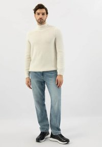 Muž v kr émovém pleteném roláku, světlomodrých džínách a černých teniskách, stojící na pozadí čistě bílé stěny.