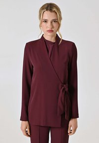 Top avvolgente bordeaux realizzato in tessuto liscio con maniche lunghe, dettaglio con fiocco laterale e vestibilità ampia, abbinato a pantaloni coordinati.