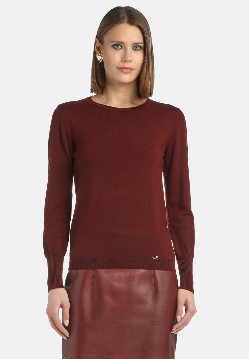 Pulover burgund fabricat dintr-un material moale, cu mâneci lungi, tiv ripsat și un accent mic cu logo în partea de jos. Asortat cu o fustă din piele.