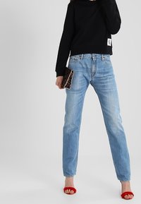 Osoba ubrana w jasnoniebieskie dżinsy, czarną bluzkę z długim rękawem, czerwone buty na obcasie z odkrytymi palcami, trzymająca kopertówkę w panterkę na szarym tle.