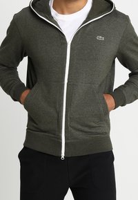 Man som bär en mörkgrön zip-up hoodie med vit dragkedja och liten krokodillogo på bröstet, med händerna i fickorna och svarta byxor.