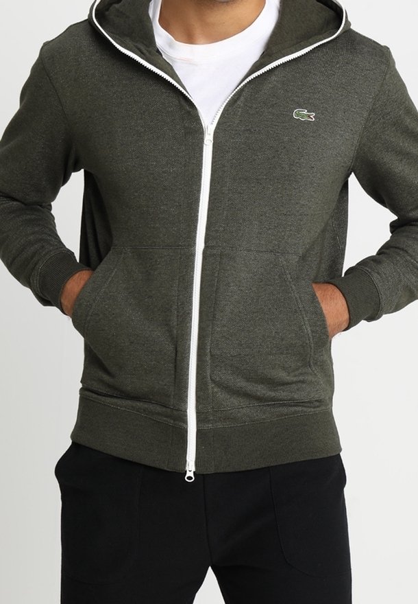 Man som bär en mörkgrön zip-up hoodie med vit dragkedja och liten krokodillogo på bröstet, med händerna i fickorna och svarta byxor.