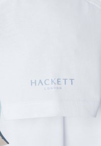 T-shirt en coton blanc avec un logo bleu "HACKETT LONDON" imprimé près du col, présentant une texture lisse et des lignes épurées.