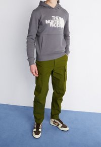 Sudadera gris con capucha y letras blancas, combinada con pantalones cargo verdes y zapatillas marrón/blancas, de pie contra un fondo claro.