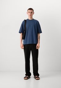 Camiseta azul de manga corta con mangas plisadas, combinada con pantalones negros y sandalias negras. El modelo lleva un peque�ño bolso bandolera negro.
