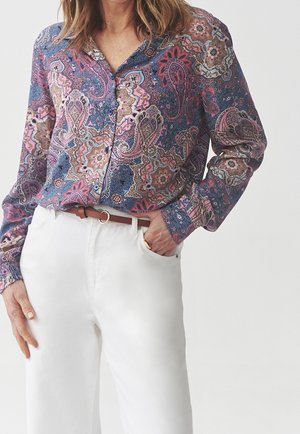 Frau trägt eine langärmelige Bluse mit Paisley-Muster in Rosa- und Blautönen, in weiße Hosen mit braunem Gürtel gesteckt.