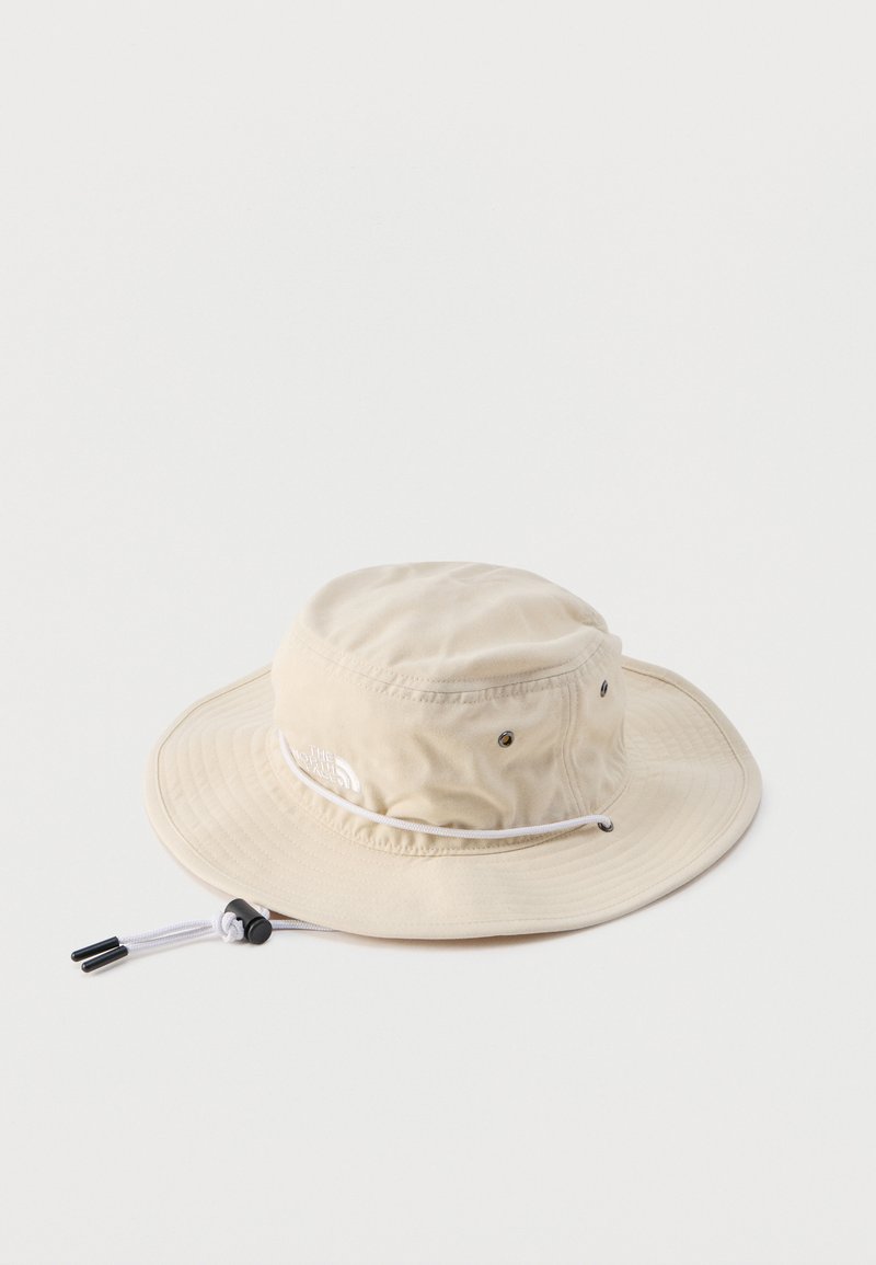 Μπεζ καπέλο με φαρδύ γείσο για τον ήλιο, με ρυθμιζόμενο λουράκι κάτω από το πιγούνι και οπές αερισμού, τοποθετημένο επίπεδα σε λευκό φόντο.