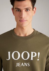 Pullover verde oliva con scollo rotondo, caratterizzato da una grande stampa bianca: "JOOP! JEANS." Tessuto liscio, materiale di peso medio, design casual.