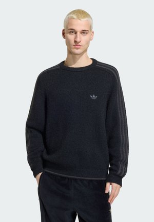 Joven con cabello rubio corto que lleva un suéter negro de Adidas con tres rayas en las mangas y el logo de Adidas en el pecho.