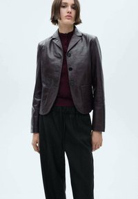 Bruine leren blazer met een getailleerd ontwerp, enige knoopsluiting, twee voorzakken en een gladde textuur. Gecombineerd met zwarte broeken.