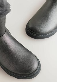 Zwarte leren laarzen met een gestructureerd oppervlak en dikke zwarte zolen, zichtbaar stiksel, en een pluizige pompon-detail op de linkerlaars tegen een lichte achtergrond.