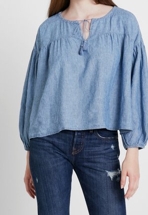 Femme portant un chemisier bleu clair à manches longues avec un empiècement froncé et un lien à pompons, assorti à un jean déchiré bleu foncé.